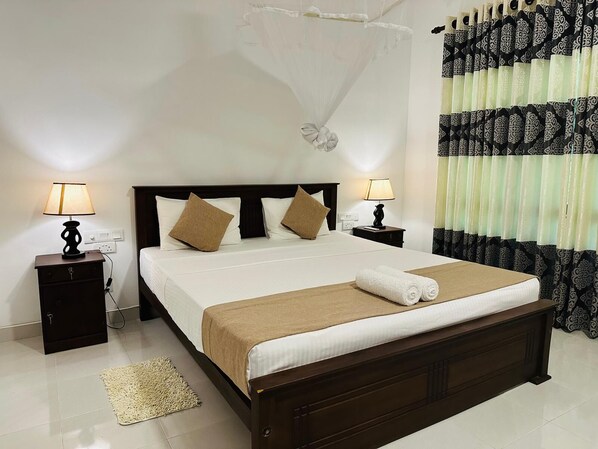 A hotel room with a large bed, two bedside tables with lamps, a bedside table with a decorative item, and a window with patterned curtains. - Sigiri Hibiscus Villa (Sigiriya)