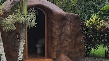 Une structure rustique, semblable à une grotte, dotée d’une porte en bois et entourée de gravier et de plantes tropicales.