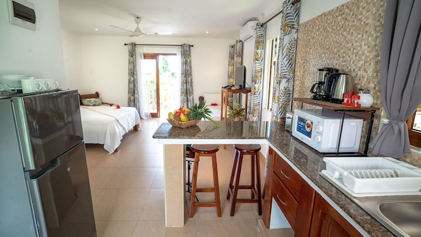 A modern kitchen with a refrigerator, microwave, and sink. There are two wooden stools and a fruit bowl on the counter. - La Maison Hibiscus (Mahé Island)
