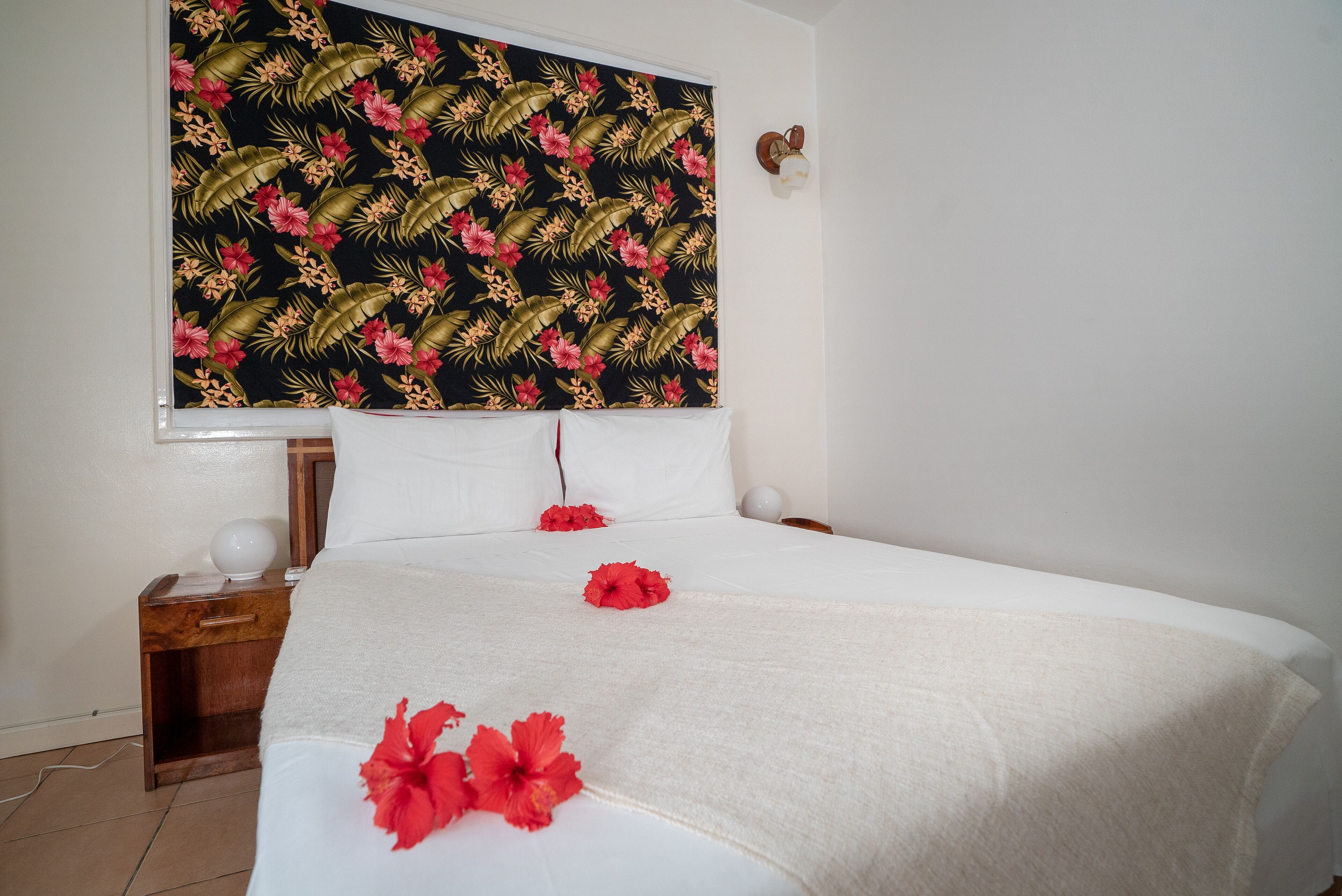 Ein ordentlich bezogenes Bett mit weißen Leinen und roten Hibiskusblüten, ein Nachttisch aus Holz mit einer Lampe sowie eine Wand mit tropischem Blatt- und Blütenmuster.
