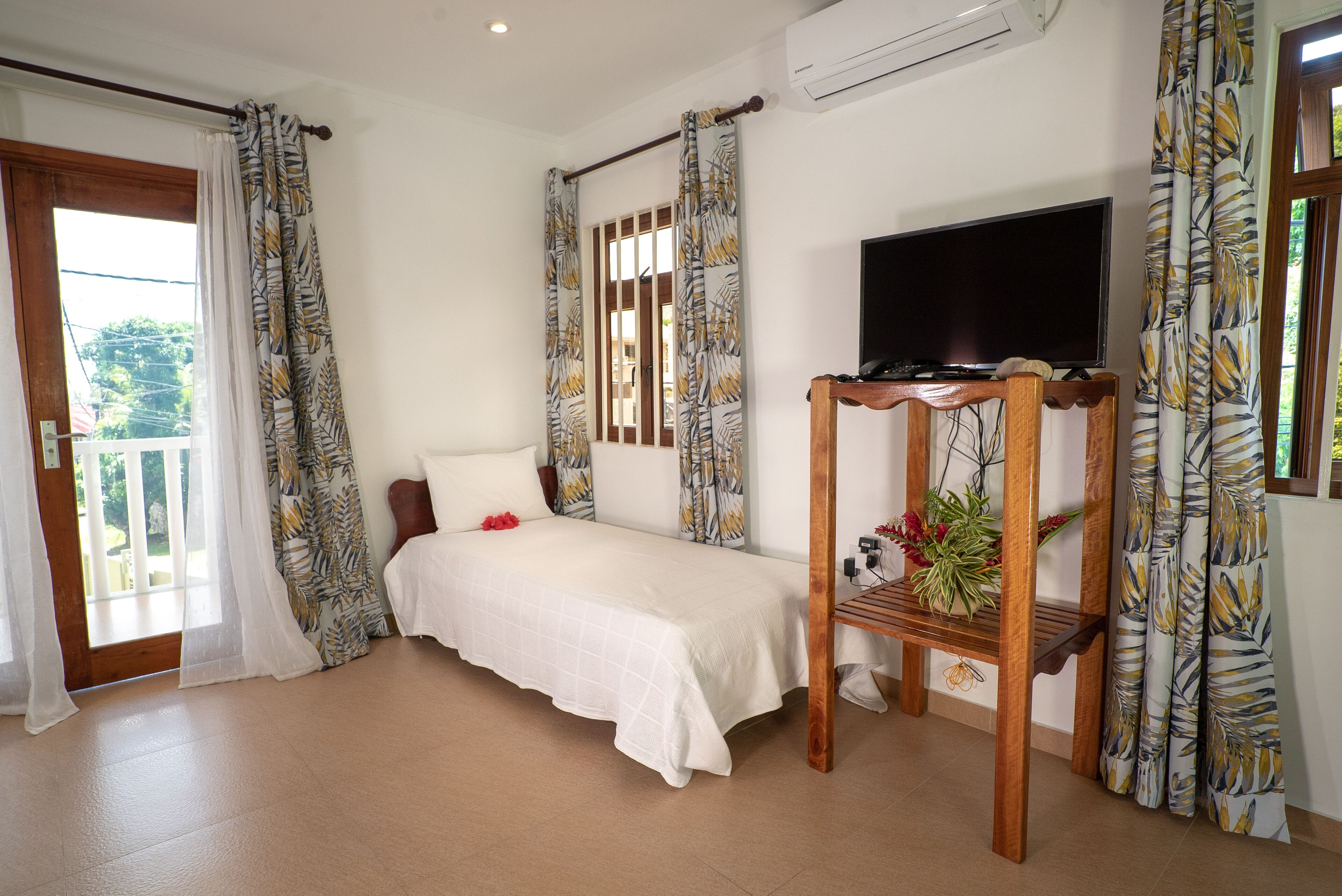 A bedroom with a bed, a TV on a wooden stand, and a balcony with curtains.