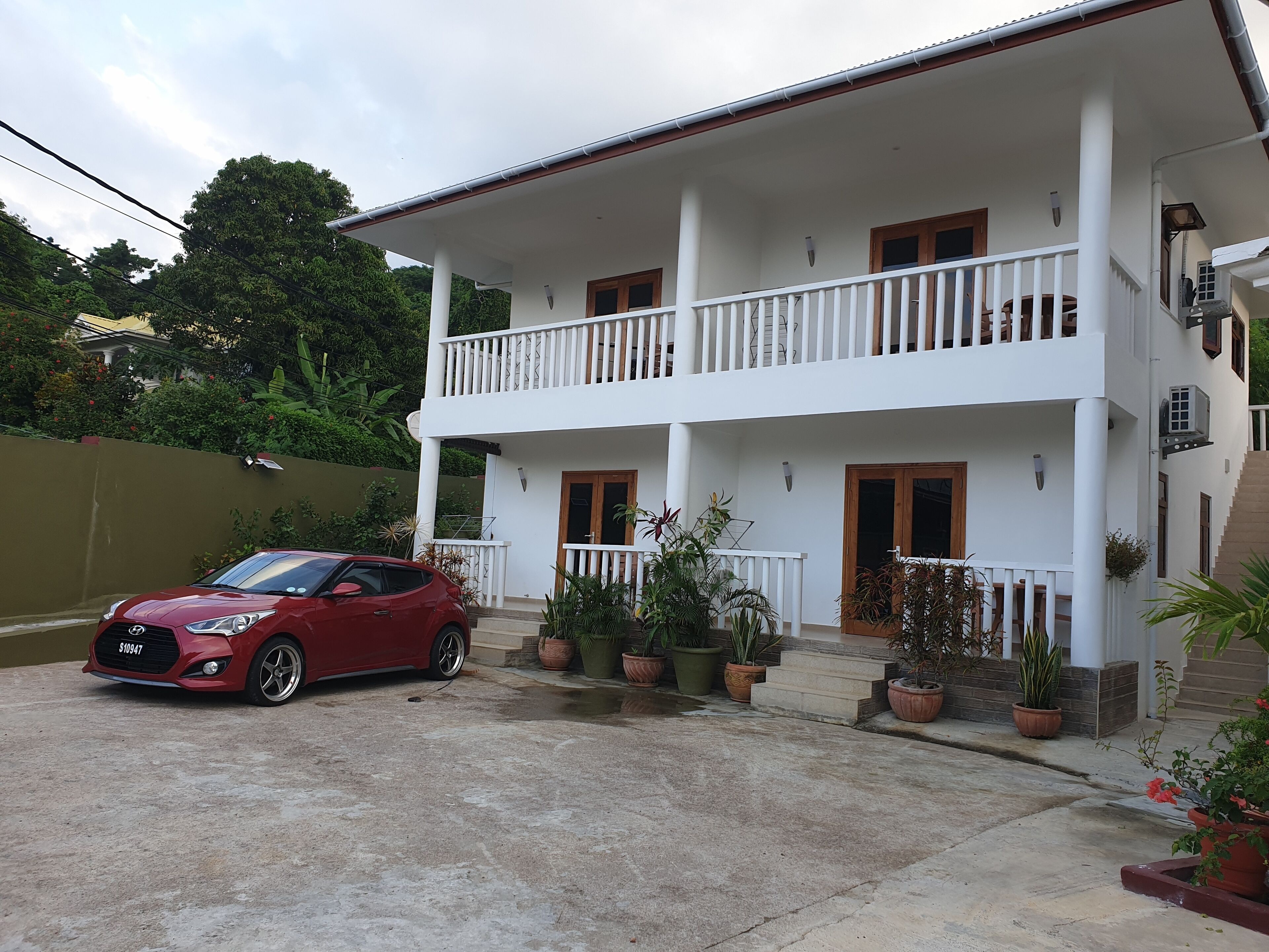 Une maison à deux étages avec un balcon, des plantes en pot et une voiture rouge garée dans l’allée.