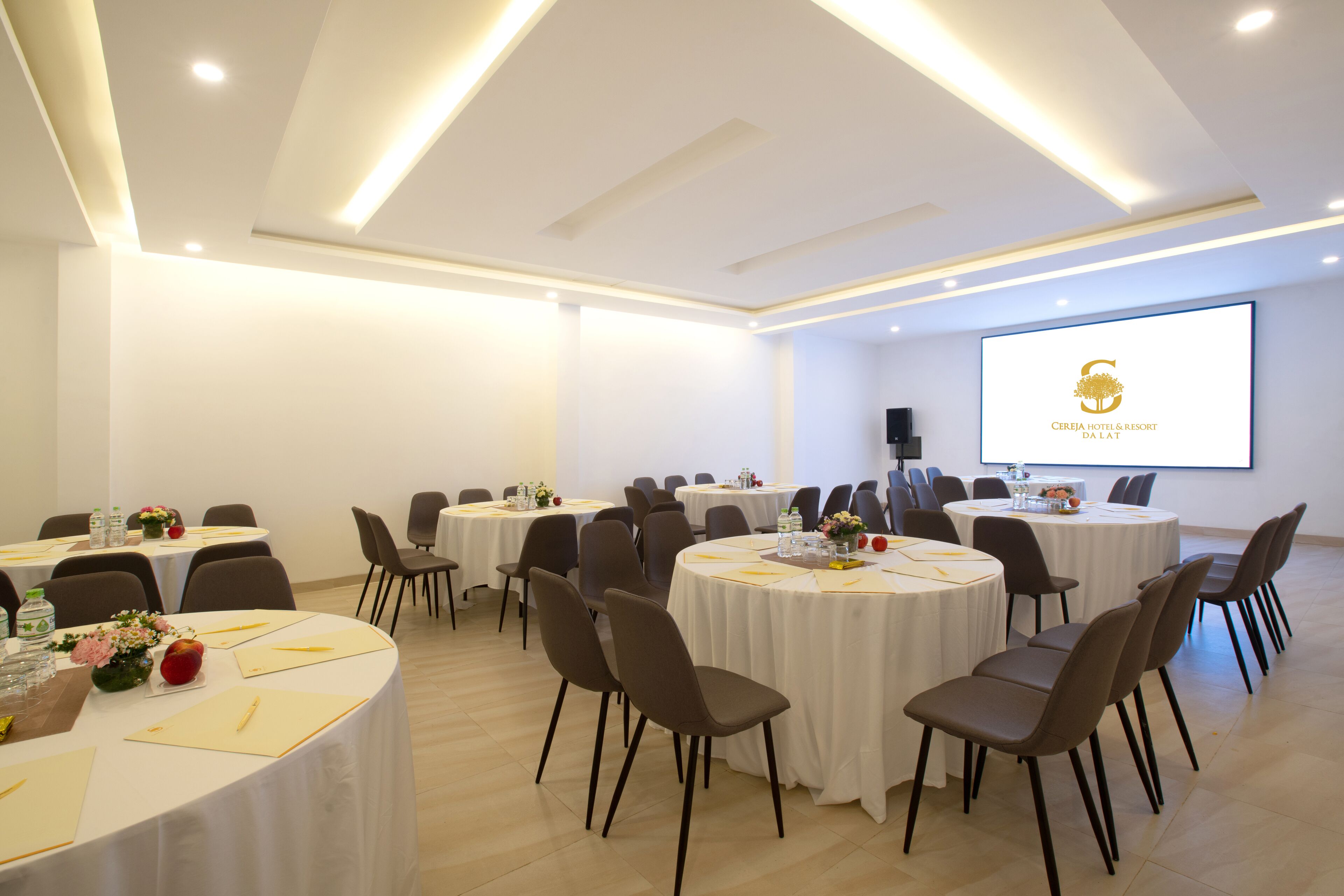 A conference room with multiple round tables, chairs, and a projector screen displaying a logo.