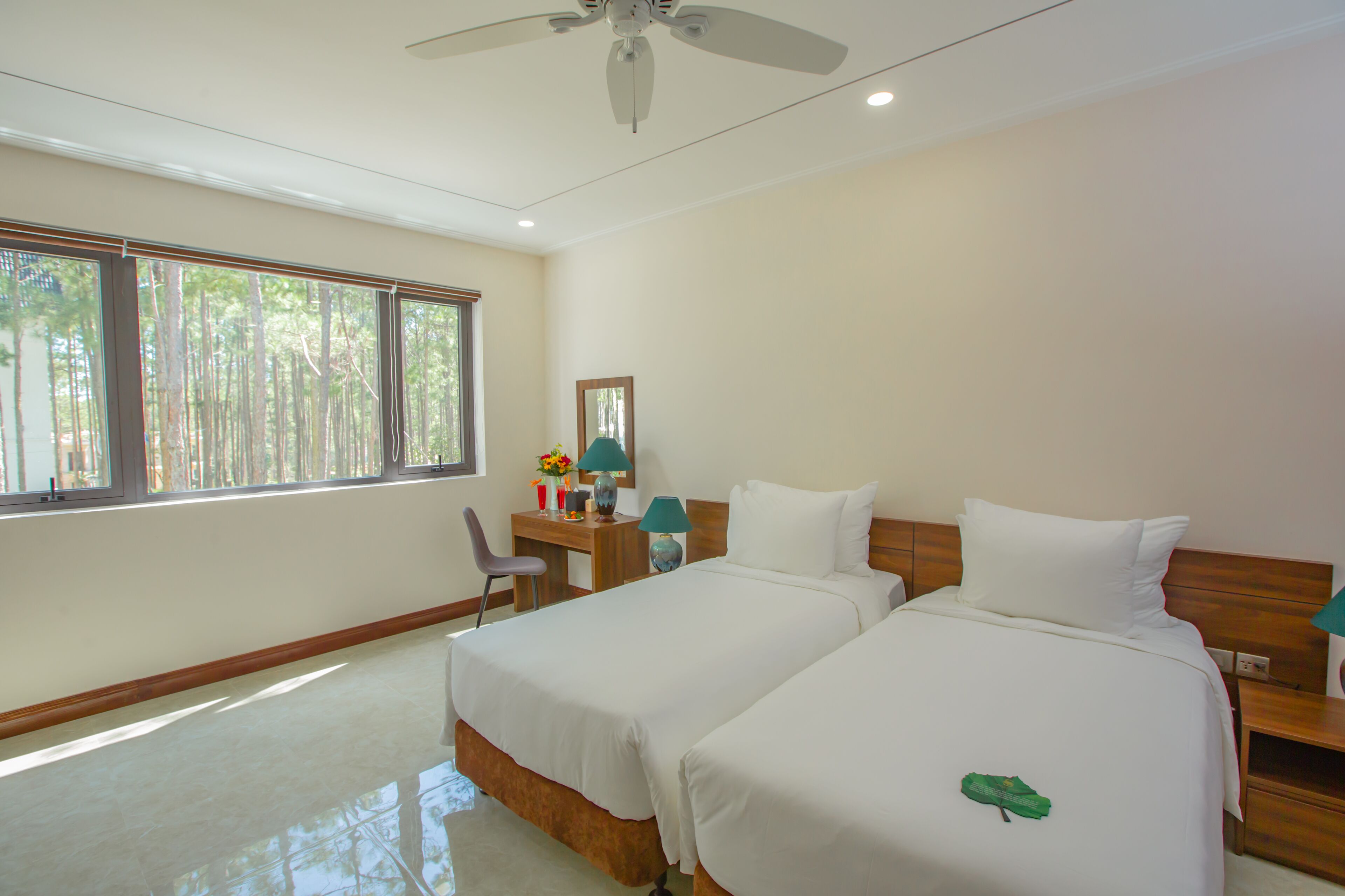 A hotel room with two beds, a desk with a chair, a ceiling fan, and large windows with a view of trees.