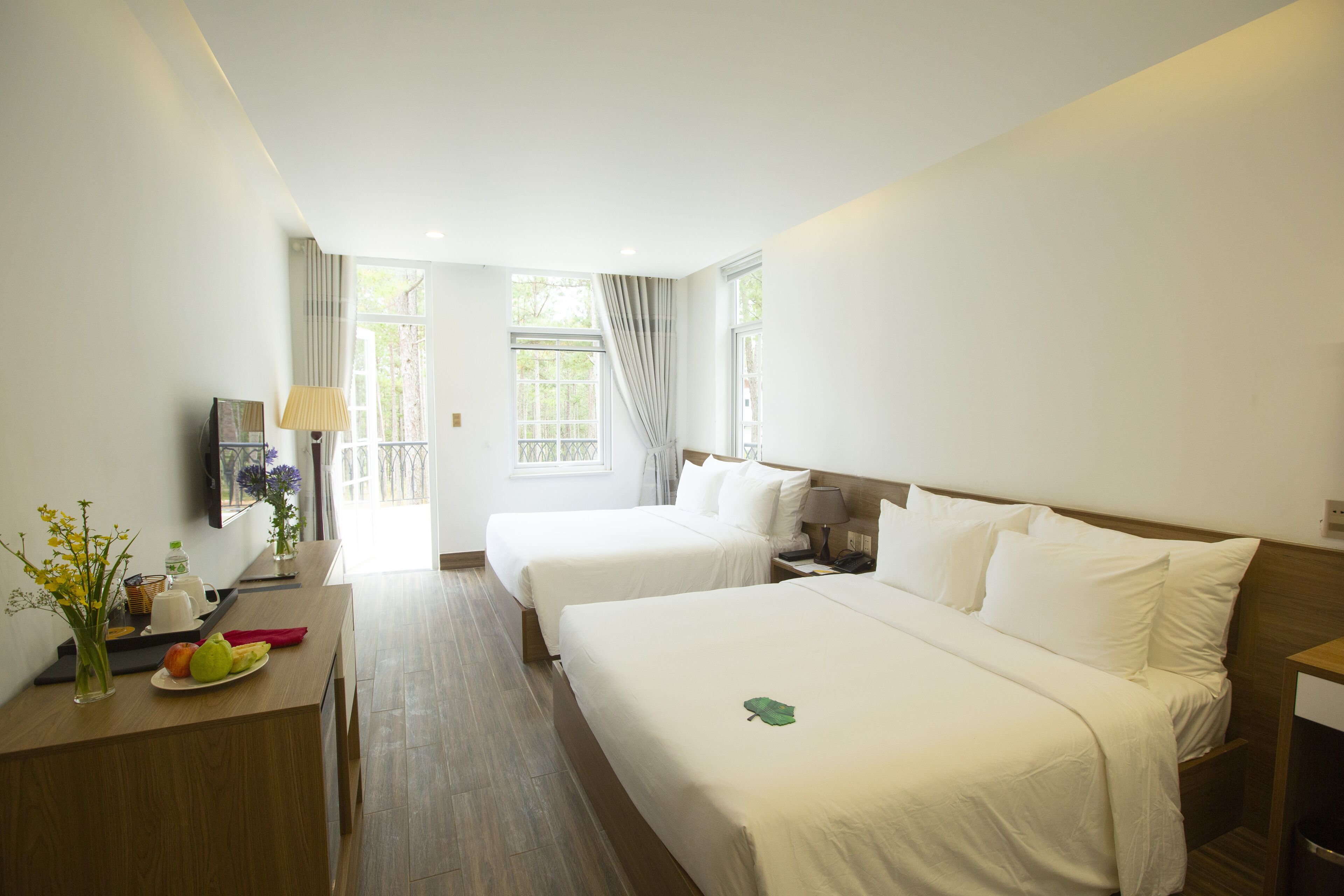 A hotel room with two beds, a desk with a lamp, a small table with a vase of flowers, and a window with curtains.