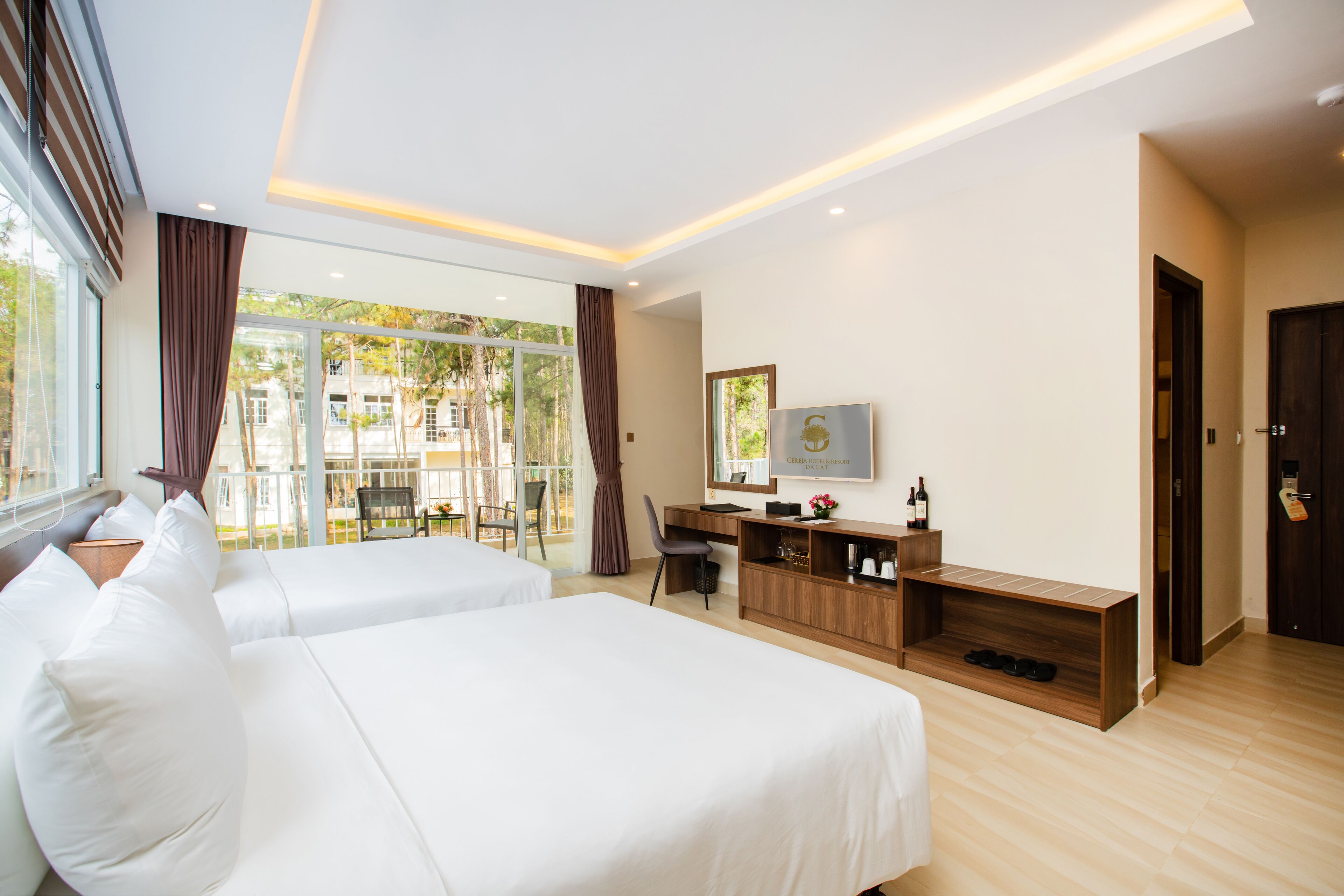 A hotel room with two beds, a desk, a TV, and a large window with a view of the outdoors.