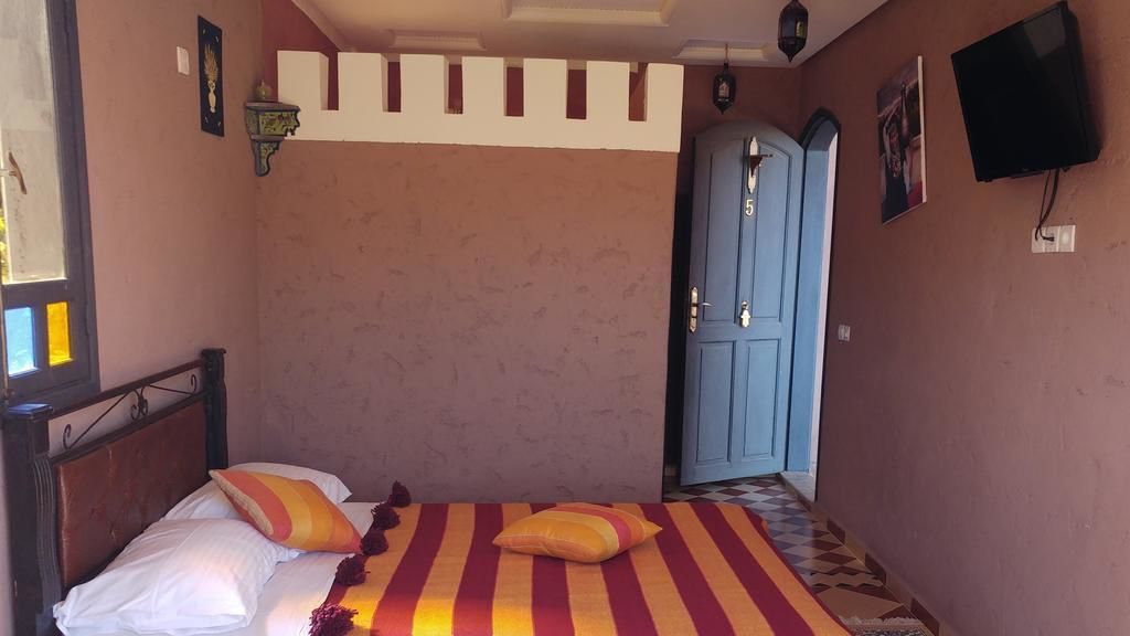 Un dormitorio con cama, almohadas, una manta a rayas, una puerta azul y un televisor montado en la pared.