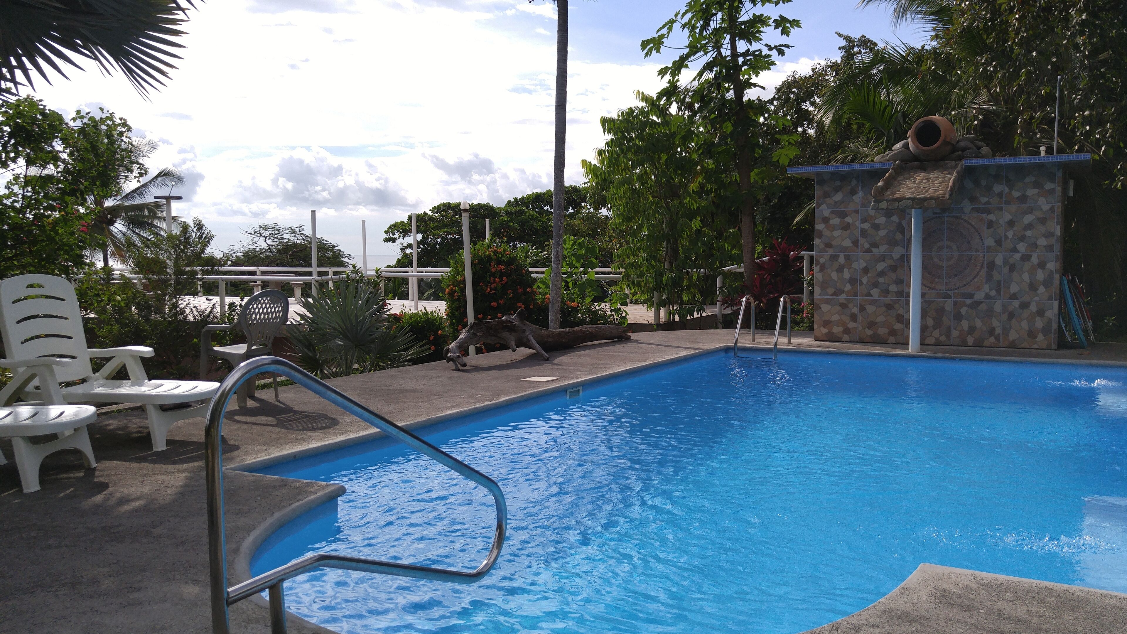 A swimming pool with a metal ladder, surrounded by white plastic chairs and a stone wall.