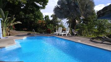 Piscina com escada de metal, rodeada por espreguiçadeiras brancas e palmeiras.