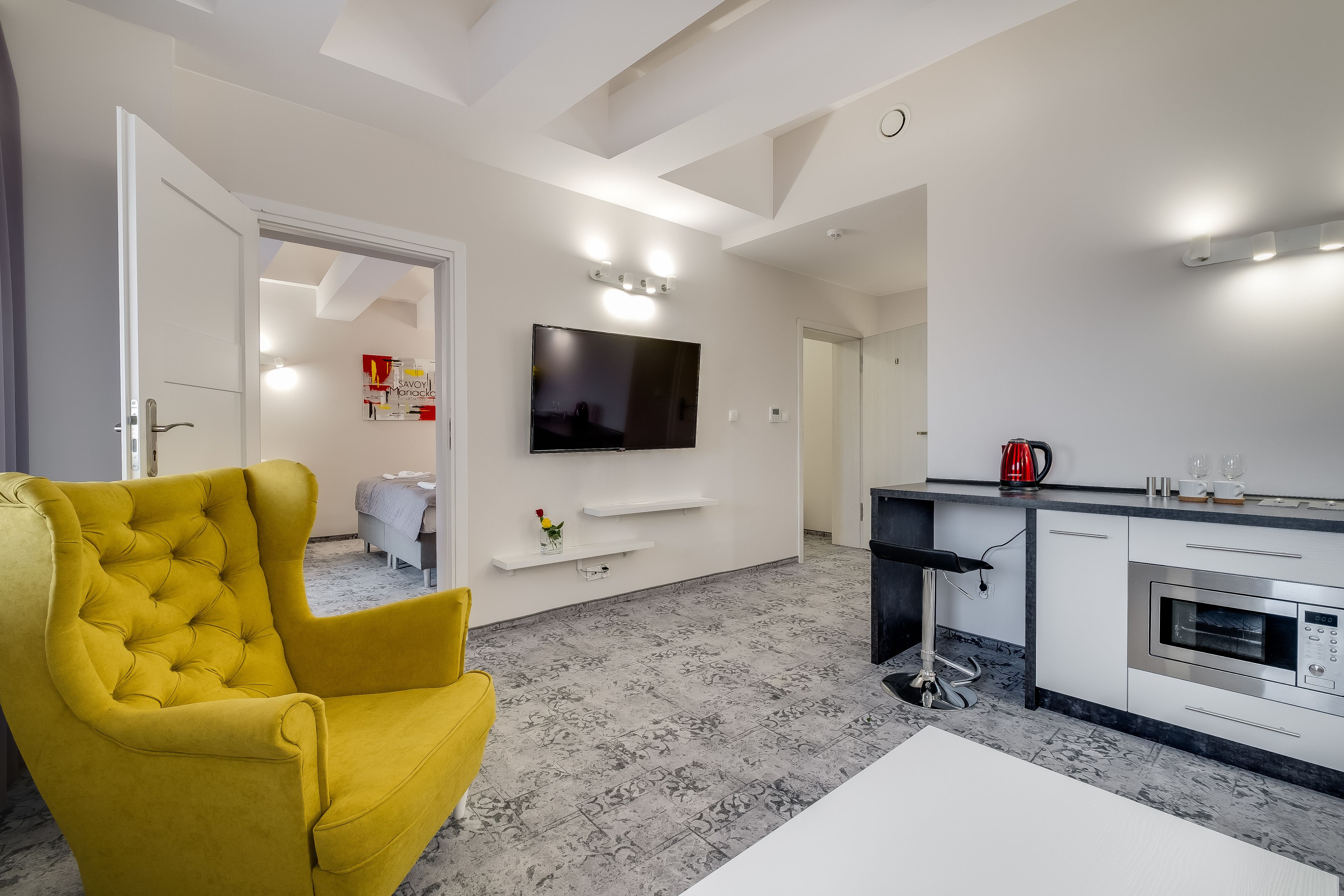 A modern living room with a yellow armchair, a flat-screen TV mounted on the wall, a kitchen area with a built-in oven, and a marble-patterned floor.
