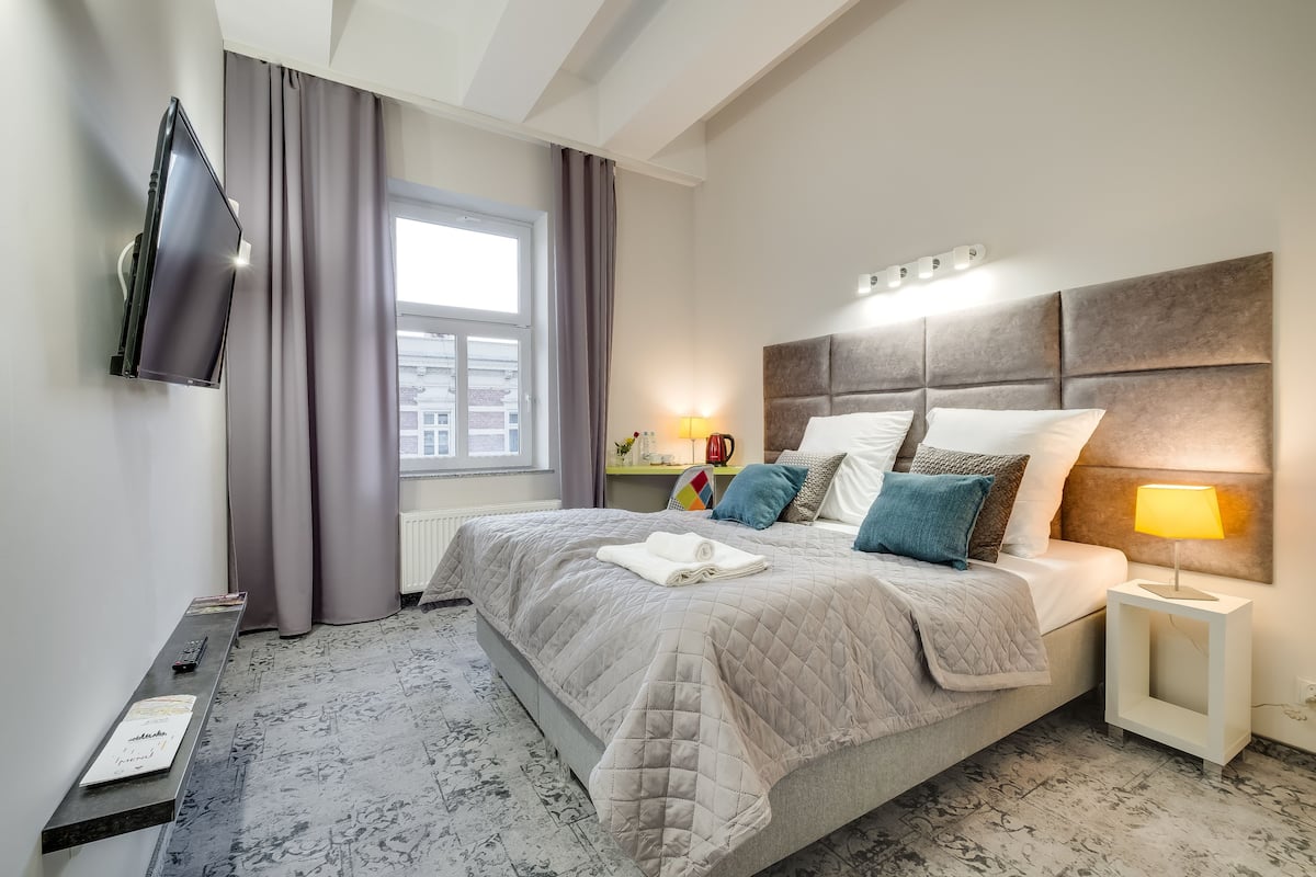 A hotel room with a bed, a TV mounted on the wall, a bedside table with a lamp, and a window with curtains.