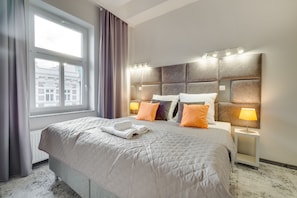A hotel room with a large bed, a headboard with lights, a window with curtains, and a nightstand with a lamp. - SAVOY Mariacka Apartments (Katowice)