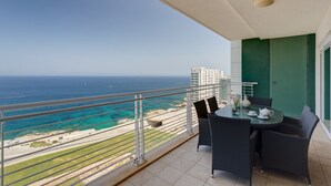 A balcony with a view of the ocean and a building across the water. There is a table with chairs and a small plant.