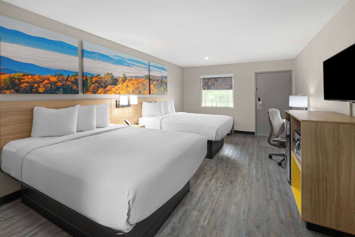 A hotel room with two beds, a desk, a chair, a TV, and a painting of autumn foliage.
