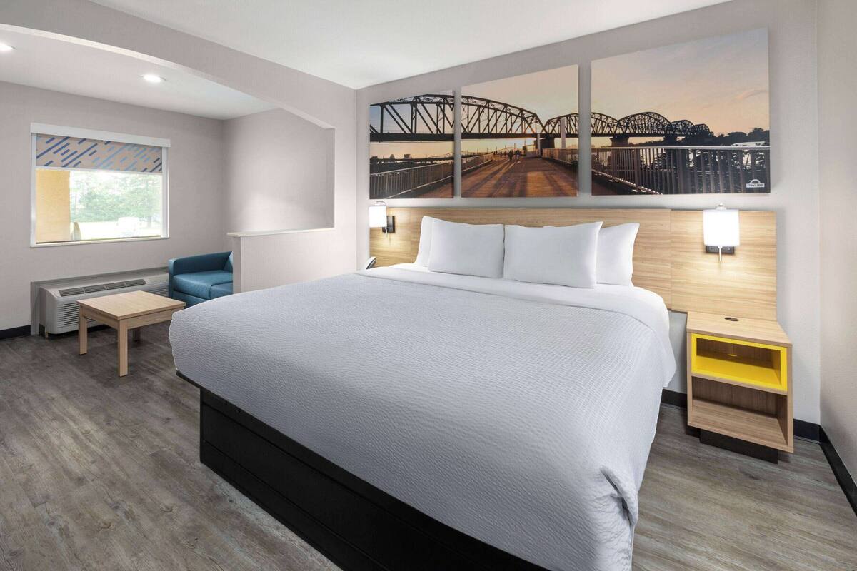 A modern hotel room with a large bed, a desk, and a chair. There is a window with blinds and a picture of a bridge on the wall.