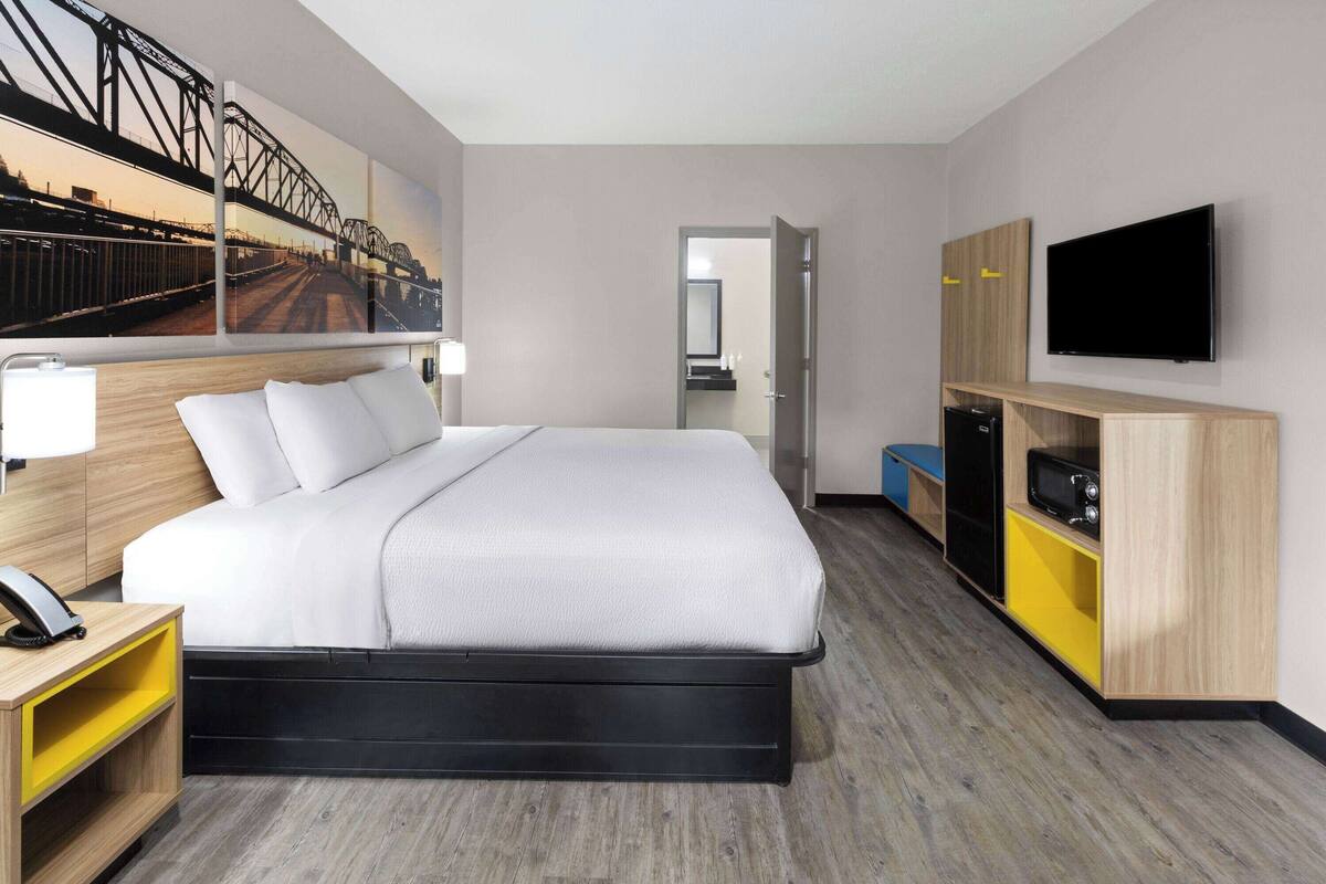 A modern hotel room with a large bed, a flat-screen TV, a wooden wardrobe, and a painting of a bridge on the wall.