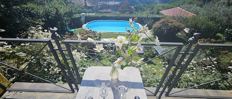Una terraza con una mesa, dos vasos y un jarrón con flores. Al fondo, una piscina rodeada de vegetación y casas.