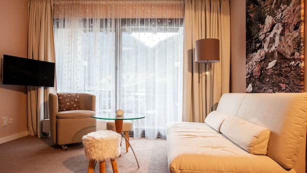 Una habitación de hotel con un sofá color beige, una mesita redonda pequeña, una silla, un taburete mullido, un televisor montado en la pared y un ventanal con cortinas traslúcidas.