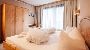 Un dormitorio con estructura de cama de madera, ropa de cama blanca, un armario de madera, un escritorio y una ventana con cortinas.