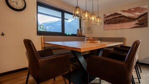 A dining area with a wooden table, four brown chairs, a clock on the wall, a window with a view of mountains, and pendant lights. - 1102 Kristall 2 (Gaschurn)