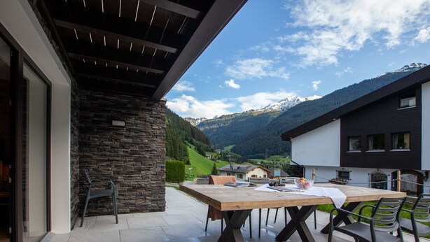 An outdoor patio with a wooden table and chairs, stone wall, and a view of mountains. - 1102 Kristall 2 (Gaschurn)