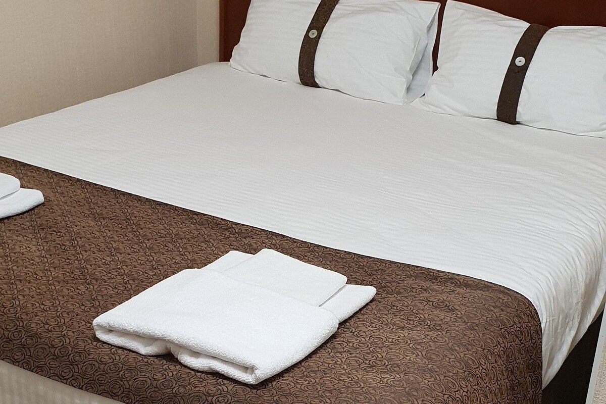 A neatly made bed with a brown and white patterned bedspread, white pillows, and a folded white towel on the bed.