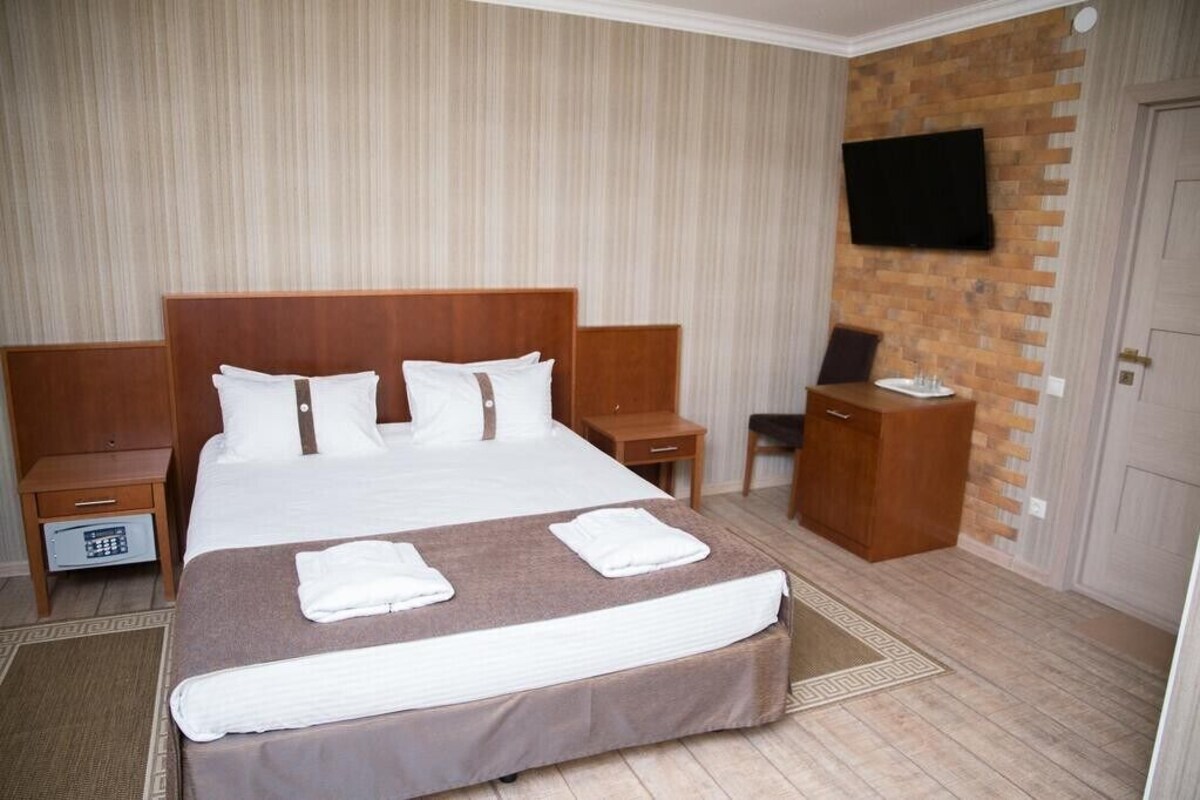 A hotel room with a large bed, wooden headboard, a flat-screen TV mounted on a brick wall, a bedside table, a nightstand, and a small wooden cabinet.