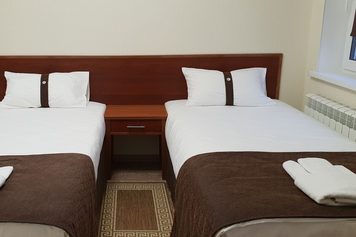A double room with two beds, each with a brown bedspread and white pillows. There is a small wooden nightstand between the beds, and a window with a brown blind to the right.