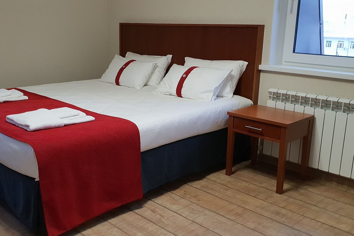 A neatly made bed with white linens and a red blanket, a wooden bedside table, and a window with a view of buildings.