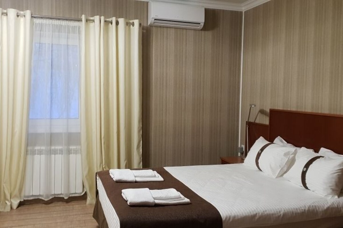 A hotel room with a bed, curtains, an air conditioning unit, and a bedside table.