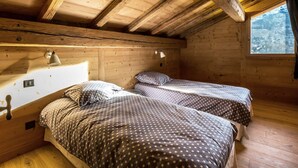 A cabin with two beds, wooden walls, a sloped ceiling, and a window with a view of a snowy landscape.