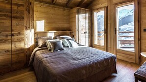 A wooden cabin bedroom with a bed, pillows, and a view of a snowy mountain.