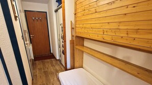Un couloir étroit avec une porte en bois, un mur blanc et un banc en bois surmonté d’un coussin blanc.