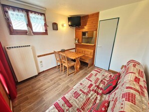 A compact room with a wooden dining table, two chairs, a bed with a patterned blanket, a television, a small kitchenette, and a window with curtains.