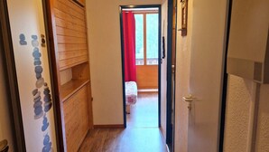 Un couloir étroit au plancher de bois mène à une pièce où se trouvent un lit et une fenêtre ornée de rideaux rouges.