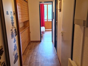 Kapea käytävä, jossa on puulattia, johtaa huoneeseen, jossa on sänky ja ikkuna, jossa on punaiset verhot.