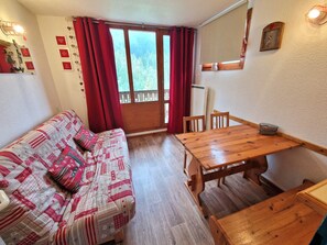 A cozy room with a wooden table and chairs, a patterned sofa, and a window with red curtains. - Studio for 4 in Valfréjus, balcony, TV, ski locker (Valfréjus)