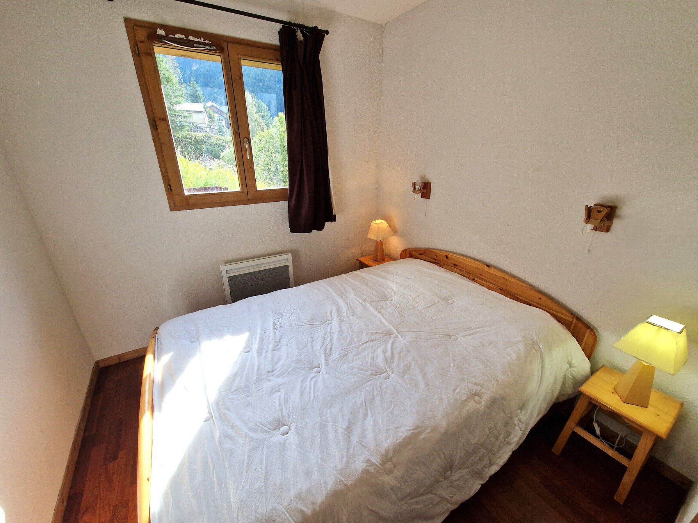 A bedroom with a bed, a window with curtains, a bedside table with a lamp, and a wall-mounted light fixture.