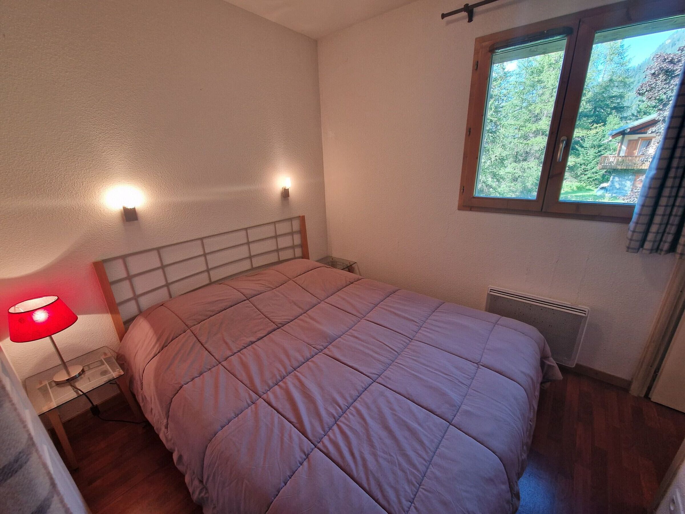 A bedroom with a bed, a nightstand, a lamp, a window, and a radiator.