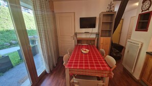Un coin repas avec une nappe à carreaux rouges, des chaises en bois, un téléviseur accroché au mur et une porte coulissante donnant sur un jardin.