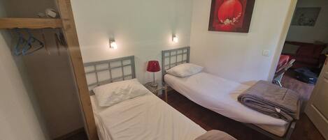 A small, compact room with two single beds, a wooden wardrobe, a red lamp, and a painting on the wall.