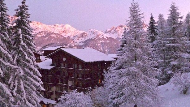 Un chalet à la façade symétrique, entouré d’arbres couverts de neige et avec des montagnes en toile de fond.