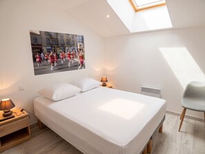 Quarto com cama, mesa de cabeceira, abajur, cadeira, radiador e claraboia.