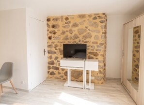 A room with a stone wall, a white desk with a flat-screen TV, a grey chair, and a wooden wardrobe.