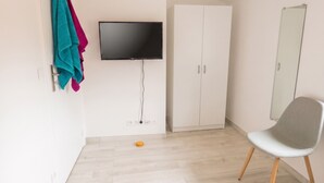 Een kamer met een aan de muur bevestigde televisie, een garderobe, een stoel en een klein oranje voorwerp op de vloer.