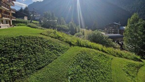 A mountainous landscape with a clear sky, a sun shining through the trees, and buildings in the foreground.