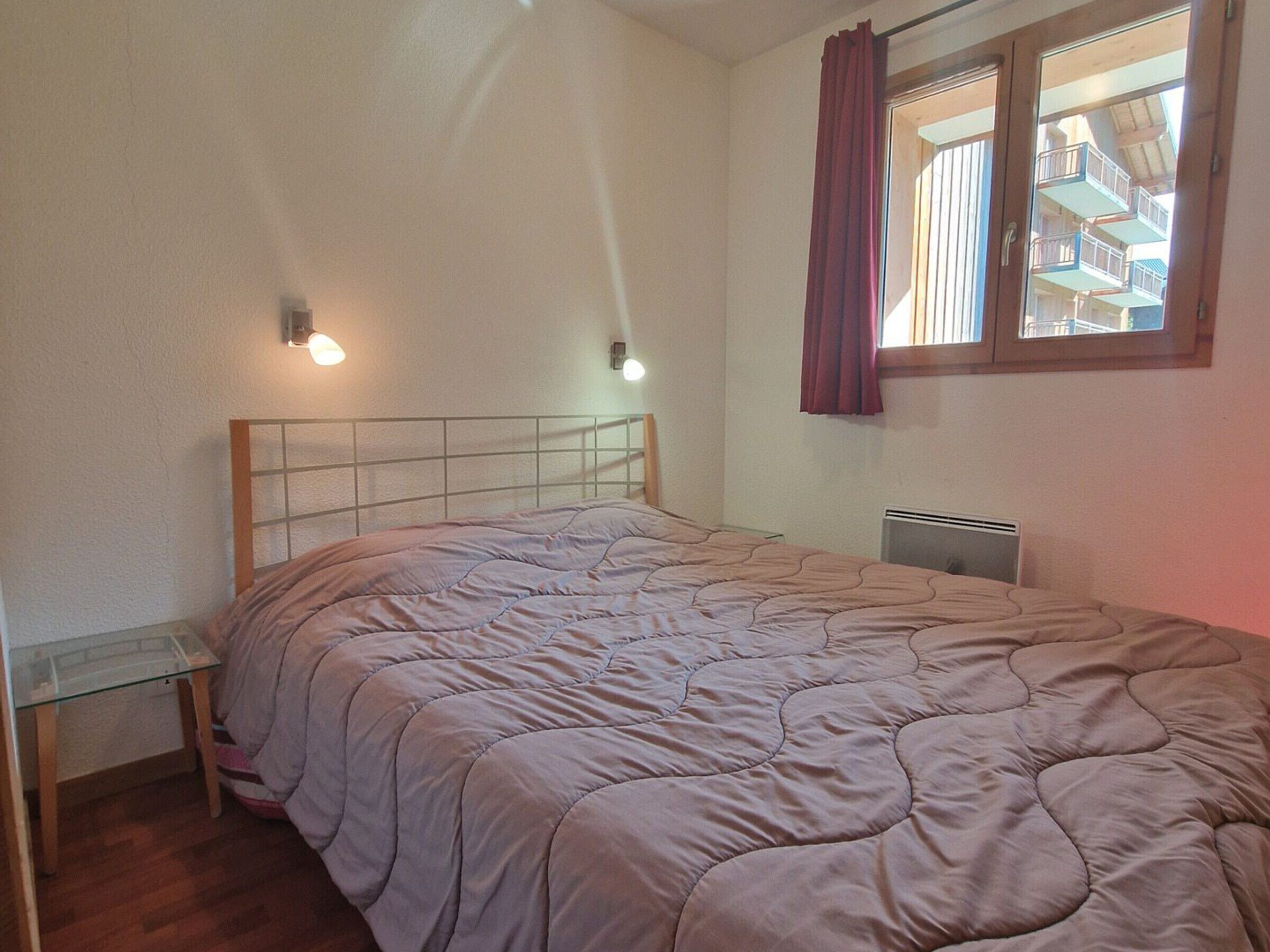 A bedroom with a bed, a glass table, a radiator, and a window with curtains.