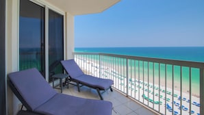 Ein Balkon mit zwei Liegestühlen, einer Glastür und Blick auf einen Strand mit Sonnenschirmen.