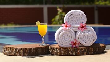Escena junto a la piscina con un vaso de zumo de naranja, toallas enrolladas y una flor.