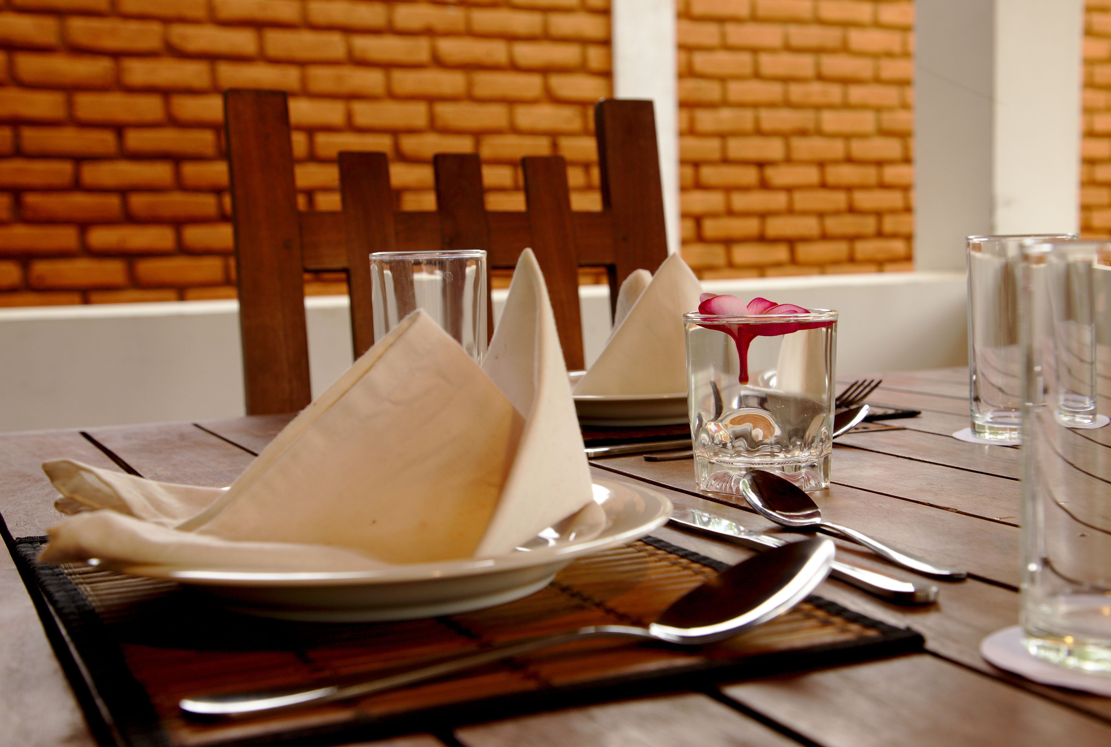 Una mesa de comedor dispuesta con platos, cubiertos y vasos, con un muro de ladrillo al fondo.